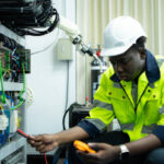 Portrait of a technician working with a digital multimeter to test the electrical system of a huge circuit board at an industrial plant using an automated hand robot.
